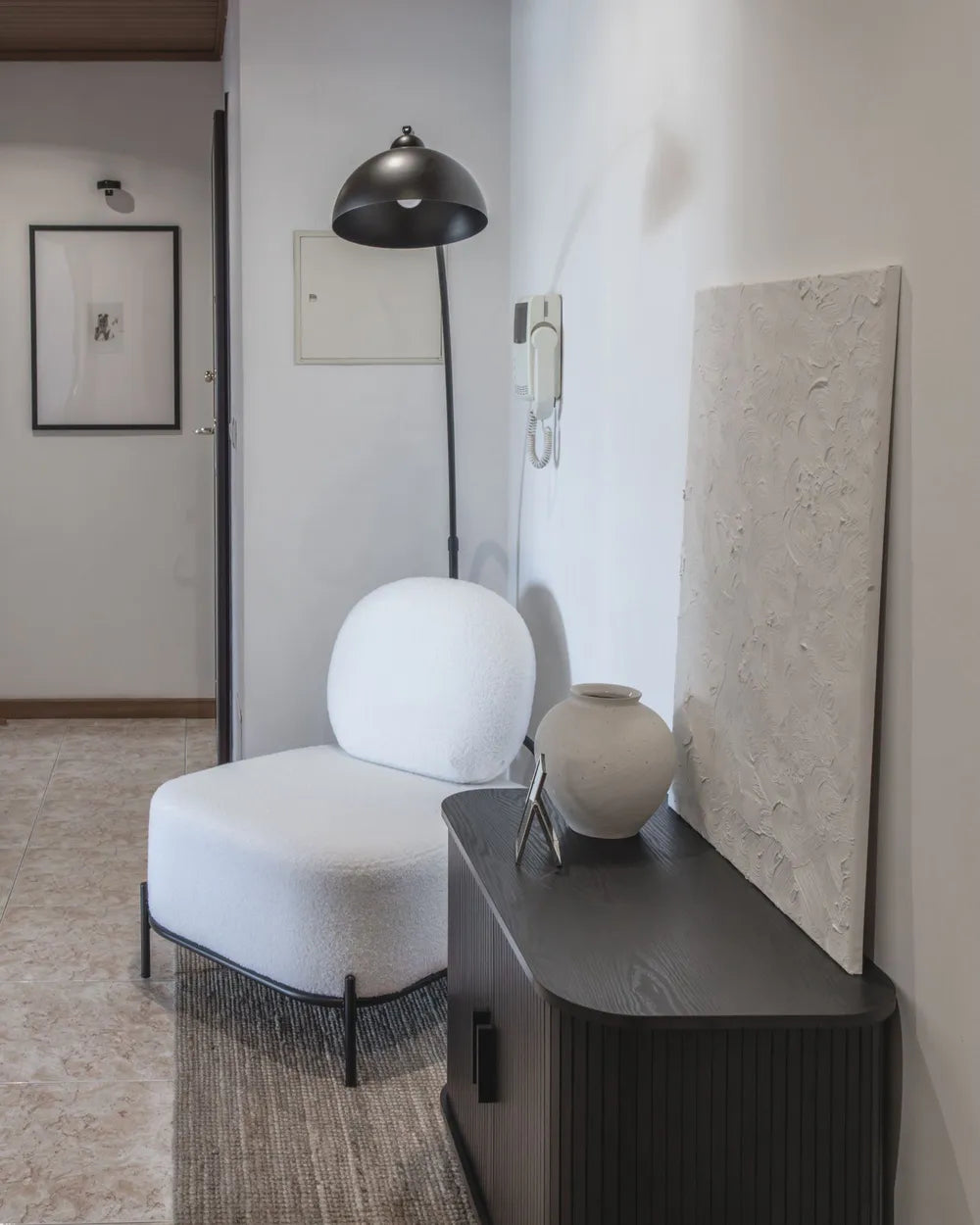 A modern white upholstered lounge chair with a rounded backrest and slim black legs, placed beside a dark wood console table in a minimalist interior. A black dome floor lamp, textured wall art, and a ceramic vase are visible in the room.
