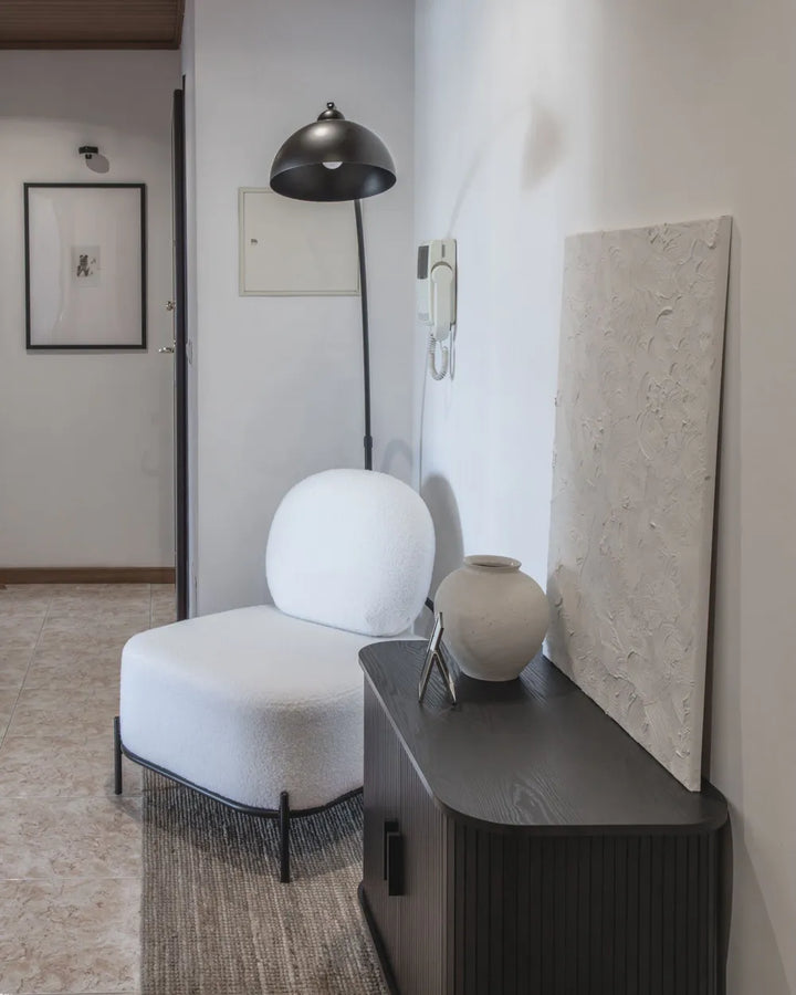 A modern white upholstered lounge chair with a rounded backrest and slim black legs, placed beside a dark wood console table in a minimalist interior. A black dome floor lamp, textured wall art, and a ceramic vase are visible in the room.