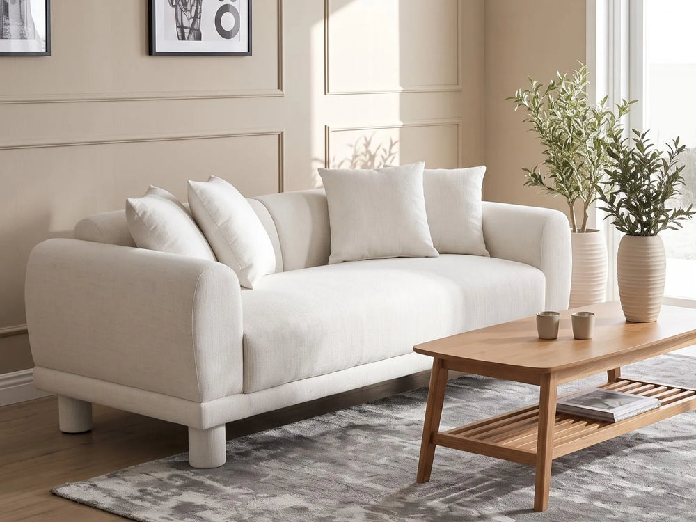 A modern cream-colored fabric loveseat sofa with matching throw pillows, styled in a bright living room next to a wooden coffee table and potted plants.