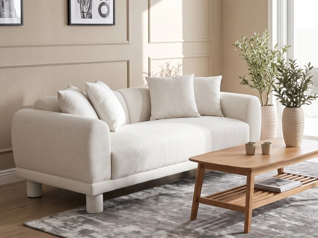 A modern cream-colored fabric loveseat sofa with matching throw pillows, styled in a bright living room next to a wooden coffee table and potted plants.
