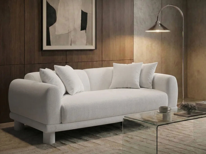 A modern white fabric sofa with rounded arms and four white throw pillows, styled in a contemporary living room setting with a glass coffee table, floor lamp, and wood-paneled wall.
