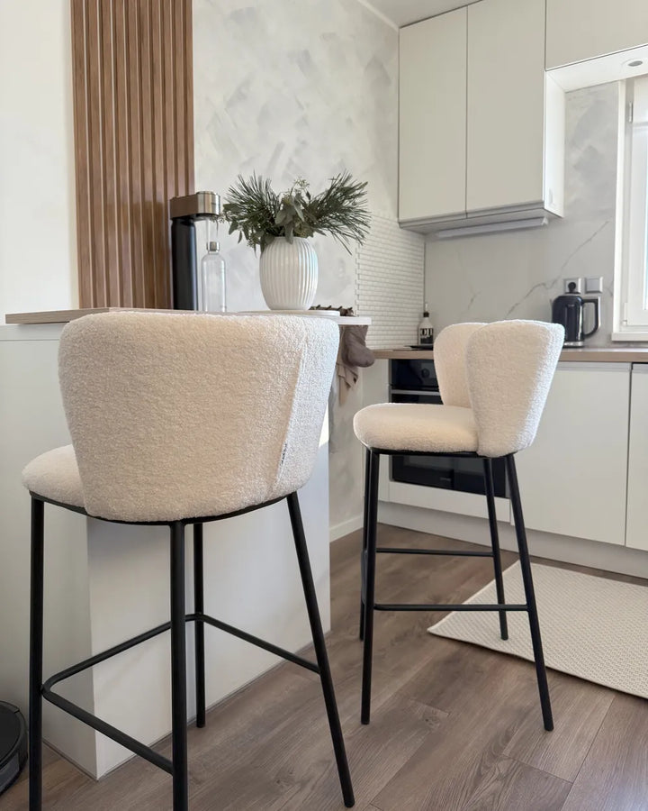 Two modern bar stools with cream bouclé upholstered seats and backs on black metal legs, positioned in a bright, contemporary kitchen setting.