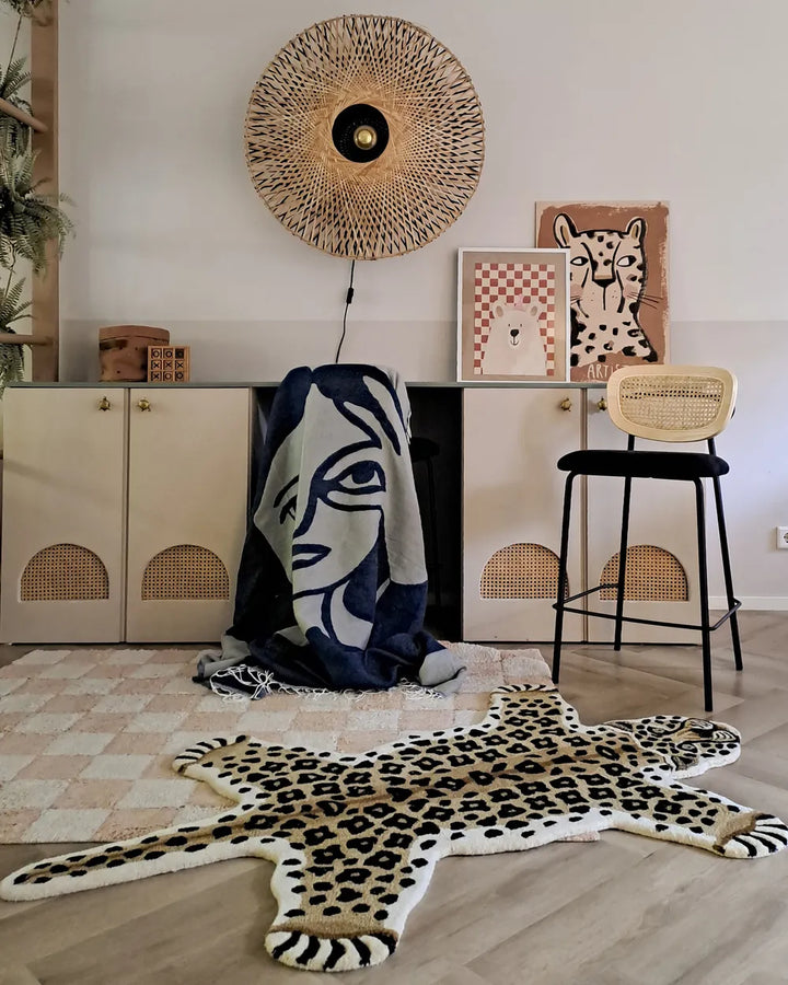 A styled interior room scene featuring a leopard-print rug on a checkered floor, a navy and cream graphic throw draped over a cabinet, a cane-topped stool, framed wall art, and a woven circular wall hanging.