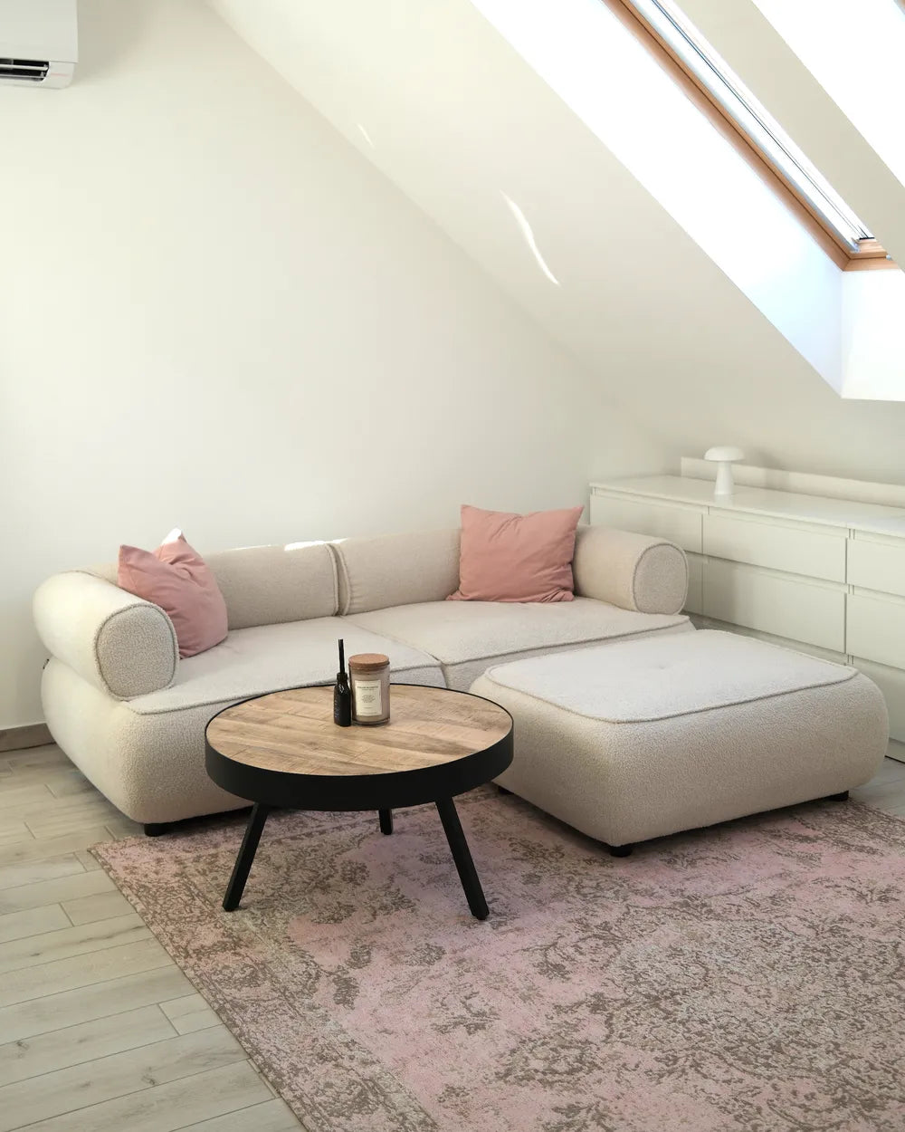 A modern light beige sectional sofa with two pink throw pillows, positioned in a bright attic room with a matching pink patterned rug and a round wooden coffee table.