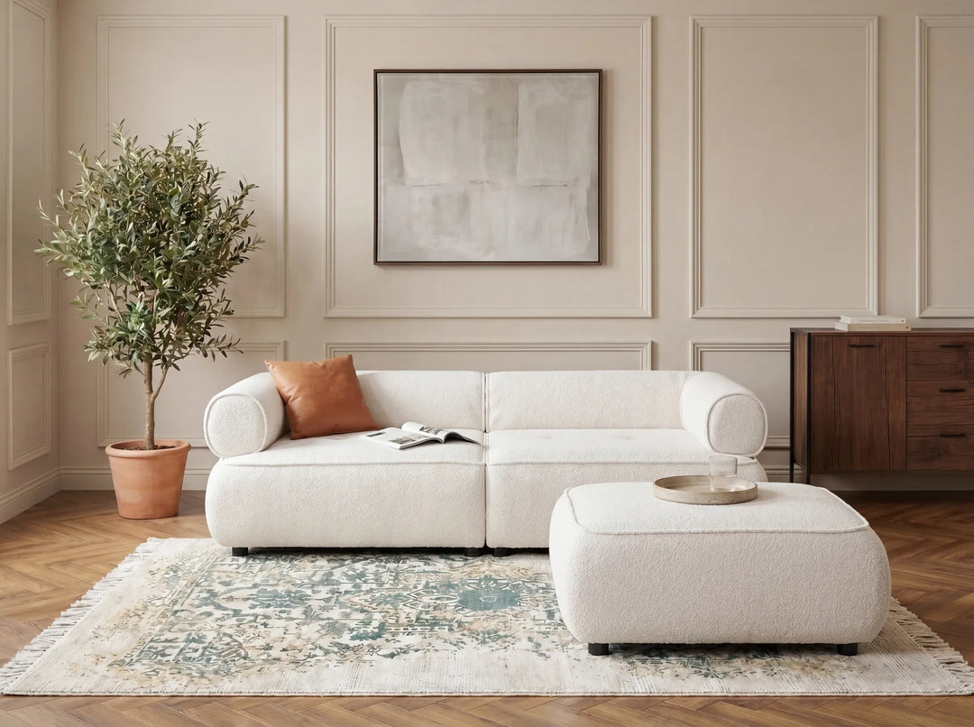 A modern white upholstered sofa with a matching ottoman in a styled living room. The room features beige paneled walls, a wooden cabinet, a potted olive tree, and a patterned rug.