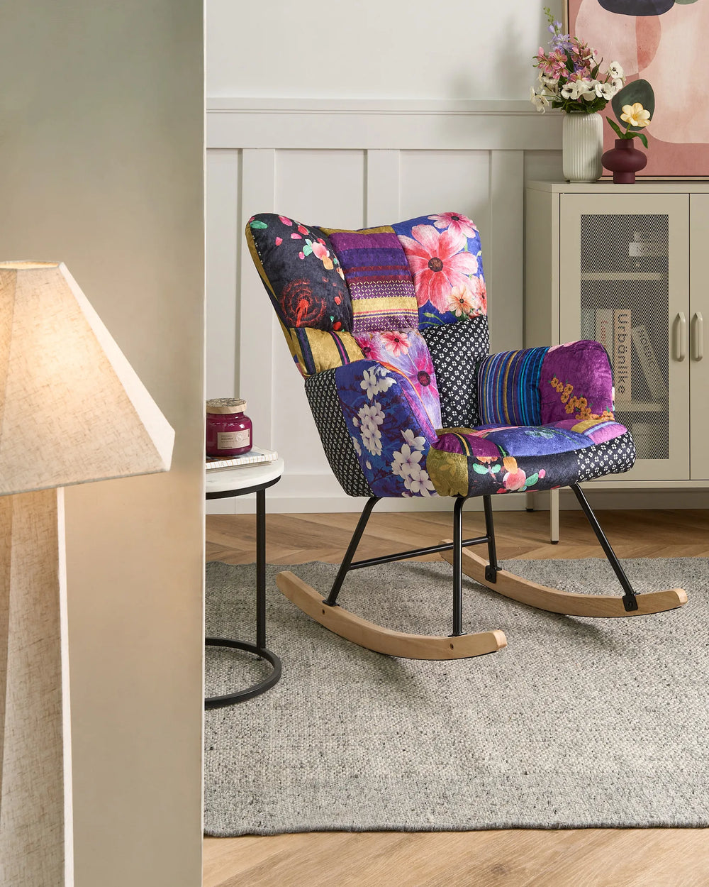 A colorful patchwork fabric rocking chair with a floral and geometric pattern, featuring black metal legs and wooden rockers, set in a styled living room next to a side table and lamp.