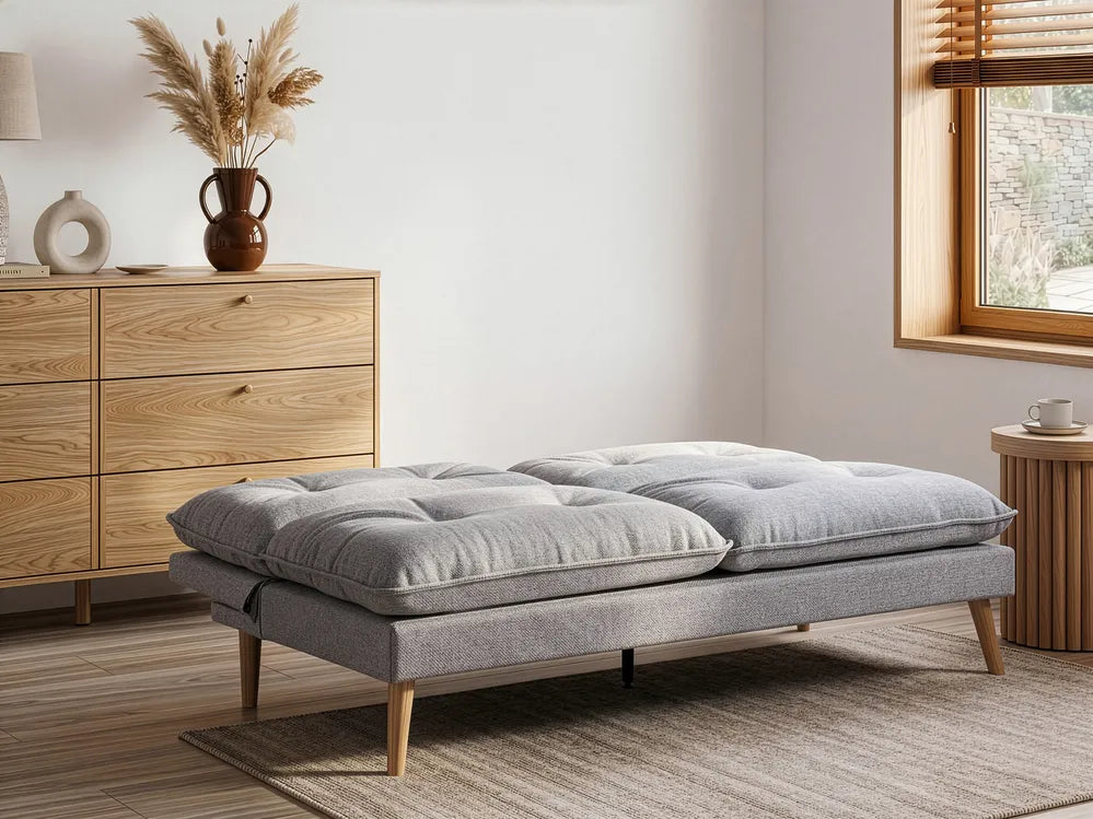 A modern gray upholstered ottoman or low bench with tufted seat cushions and tapered wooden legs, shown in a styled room with a wood dresser, rug, window, and side table.