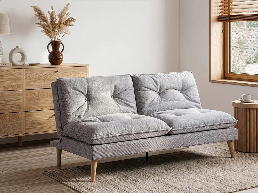 A light gray upholstered futon sofa with tufted back cushions and tapered wooden legs, shown in a styled living room setting with a wooden dresser, rug, and window.