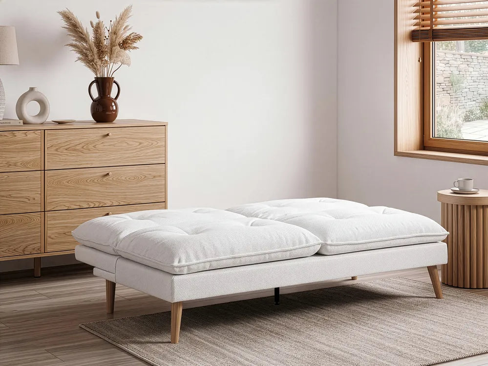 A modern upholstered ottoman or daybed with two large tufted white cushions and light wood legs, shown in a styled room with a wooden dresser, rug, window, and side table.