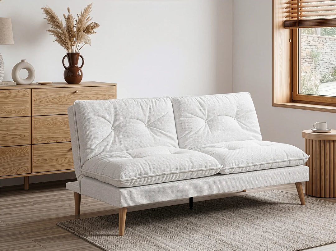 A modern two-seat sofa in off-white fabric with tufted back cushions and tapered wooden legs, styled in a bright, minimalist room with a light wood dresser, window, and rug.
