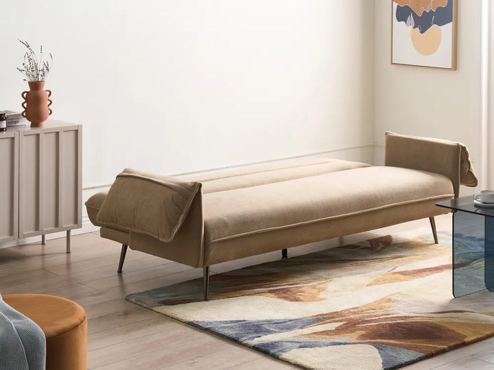 A beige upholstered daybed or chaise lounge with slim metal legs, styled in a modern living room setting with a patterned rug, side cabinet, and abstract wall art.
