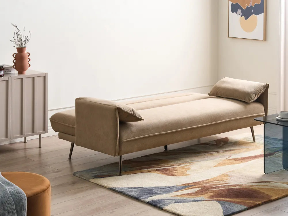 A beige upholstered daybed or chaise lounge with slim metal legs, shown in a styled living room setting with a patterned rug, side cabinet, and wall art.