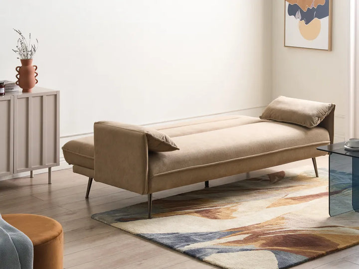 A beige upholstered daybed or chaise lounge with slim metal legs, shown in a styled living room setting with a patterned rug, side cabinet, and wall art.