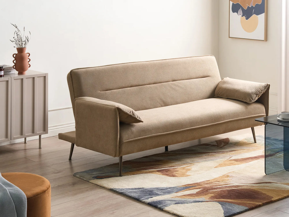 A beige upholstered sofa with slim metal legs, shown in a styled living room setting with a patterned rug, sideboard, and abstract wall art.