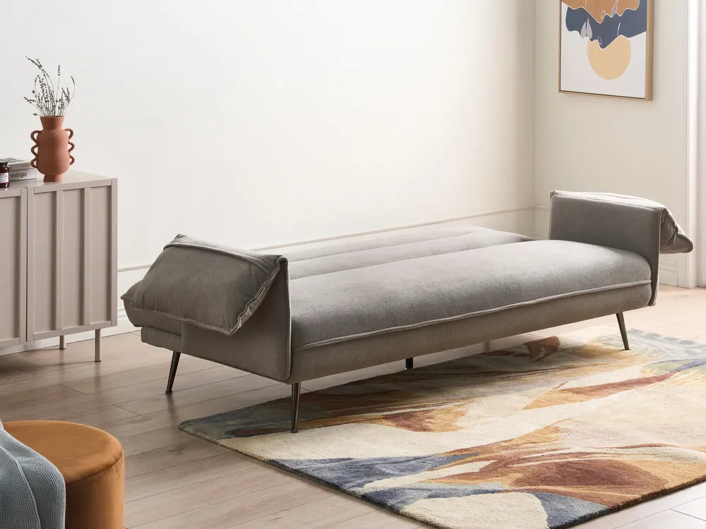 A modern gray fabric daybed with tapered black metal legs, styled in a minimalist room with a patterned rug, sideboard, and abstract wall art.