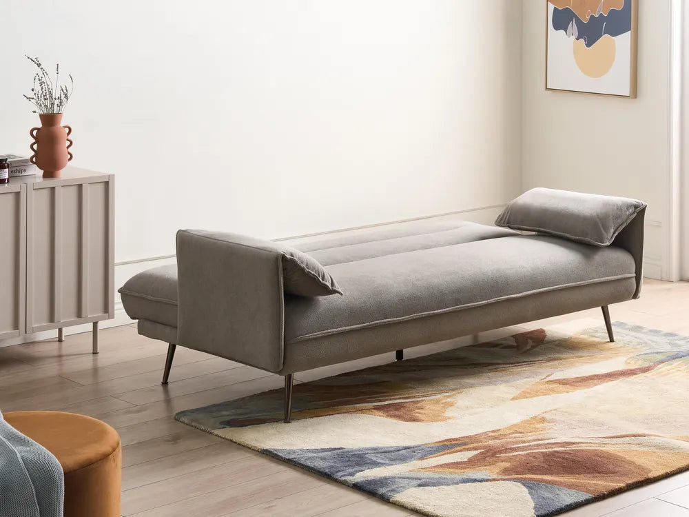 A modern gray fabric daybed with slim metal legs, styled in a minimalist room with a patterned rug, a sideboard, and abstract wall art.