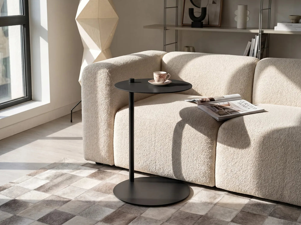 A modern black metal side table with a round top and circular base, styled next to a textured cream boucle sofa in a sunlit living room.
