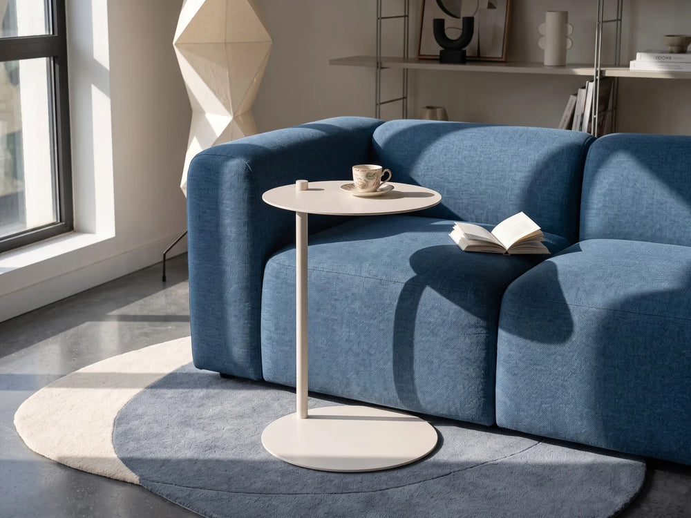 A modern blue fabric sofa in a bright living room, styled with a small round white side table, an open book, and a cup on the seat. The room features a blue rug, a geometric floor lamp, and a window casting natural light.