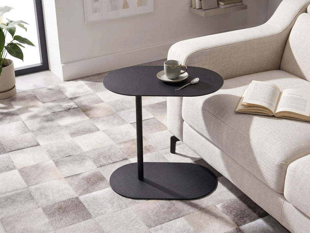 A modern black oval side table with a single pedestal leg stands beside a light beige sofa in a bright living room. A cup and saucer with a spoon sit on the tabletop, and an open book rests on the sofa.