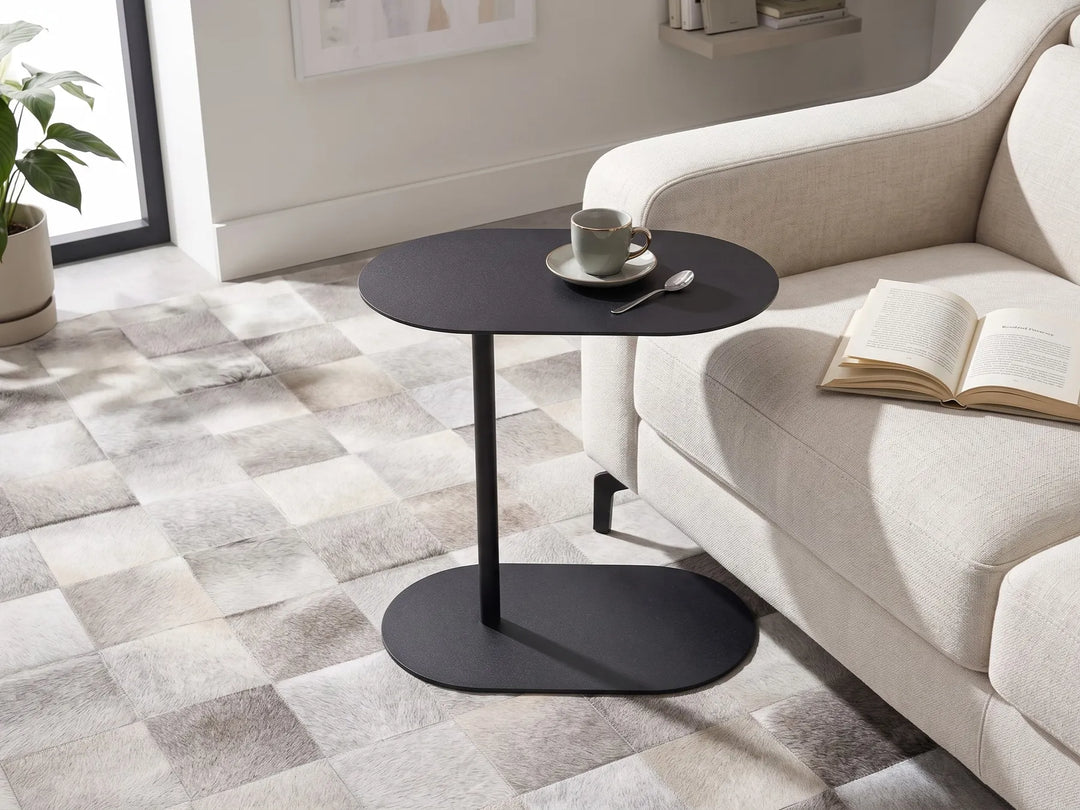 A modern black oval side table with a single pedestal leg stands beside a light beige sofa in a bright living room. A cup and saucer with a spoon sit on the tabletop, and an open book rests on the sofa.