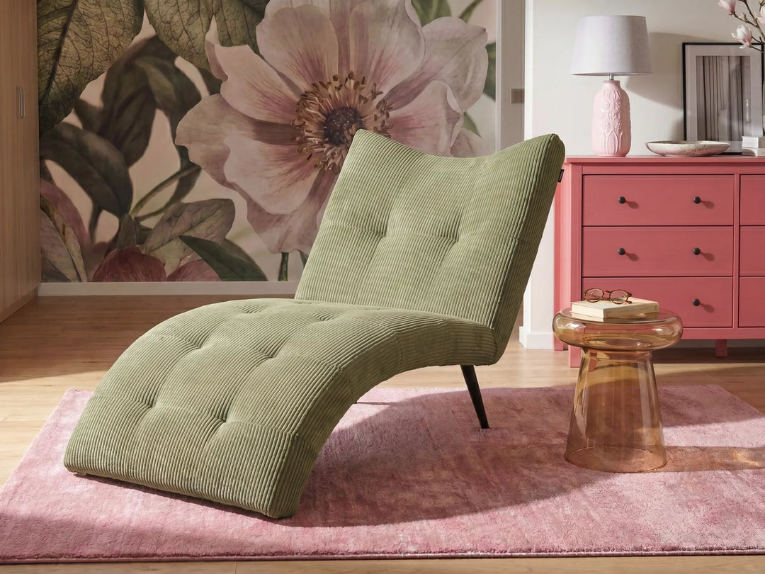 A modern olive green corduroy chaise lounge chair with tufted detailing, styled on a pink rug next to a pink side table in a room with a floral wallpaper mural and a pink dresser.