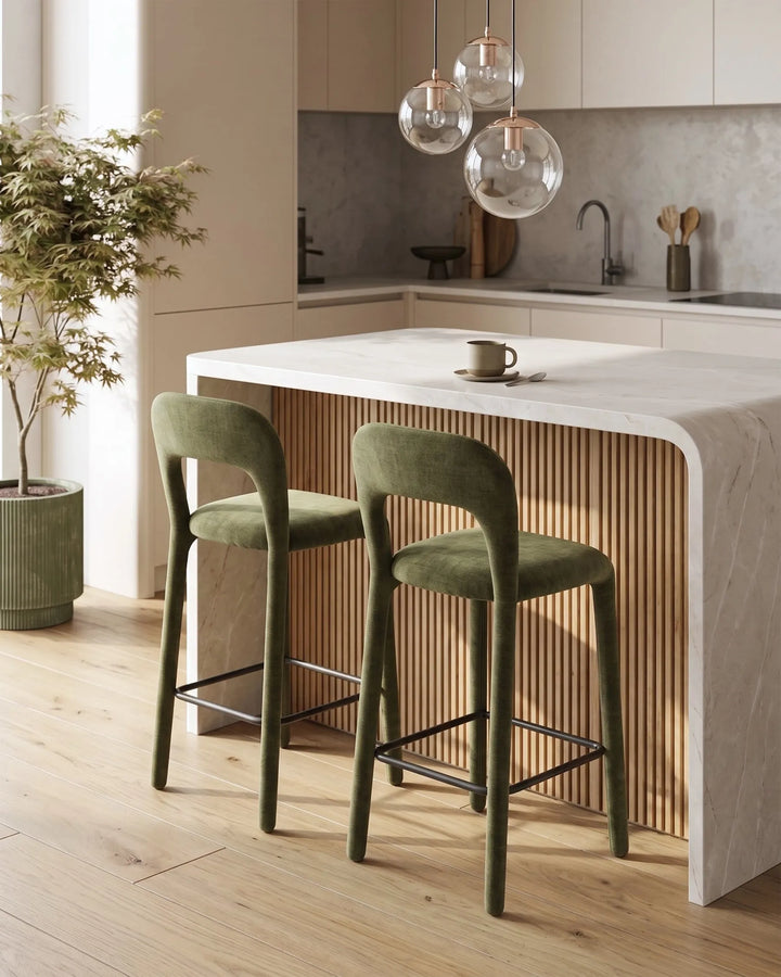 Two green upholstered bar stools with black metal legs positioned at a curved white kitchen island with a slatted wood front, in a modern kitchen setting.