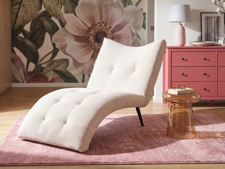 A cream-colored corduroy chaise lounge chair with tufted detailing, styled on a pink rug next to a pink side table in a room with a floral wallpaper background.