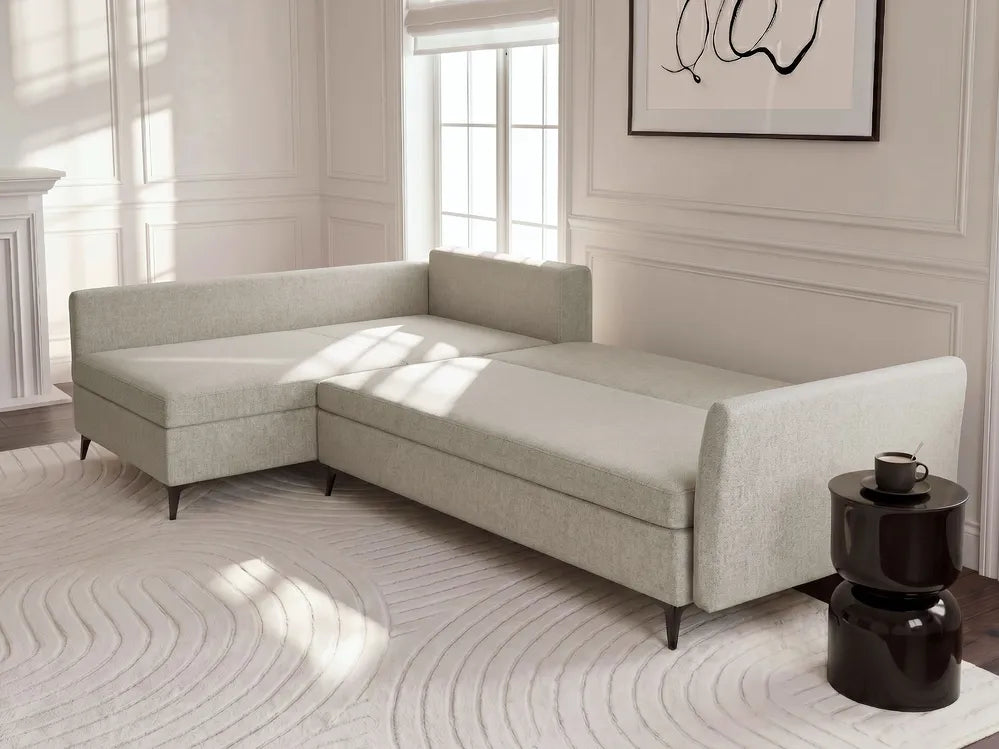 A light beige upholstered sectional sofa with a chaise lounge section, styled in a bright, modern room with a patterned rug and a dark side table.