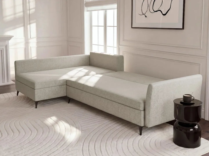 A light beige upholstered sectional sofa with a chaise lounge section, styled in a bright, modern room with a patterned rug and a dark side table.