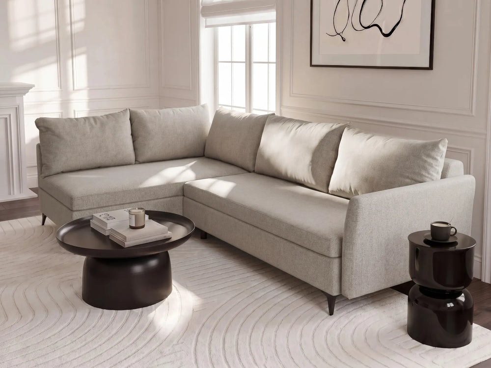 A light beige upholstered sectional sofa with matching throw pillows in a bright, modern living room. The sofa is styled with a round black coffee table and a small black side table on a white patterned rug.