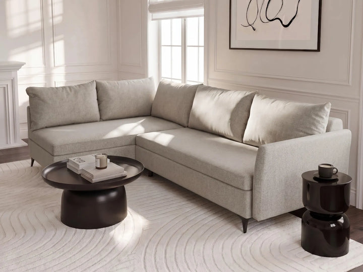 A light beige upholstered sectional sofa with matching throw pillows in a bright, modern living room. The sofa is styled with a round black coffee table and a small black side table on a white patterned rug.