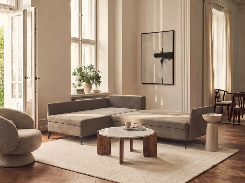 A modern L-shaped sectional sofa upholstered in a textured taupe fabric, styled in a bright, elegant living room with warm beige walls, a marble-top coffee table, and a large cream rug.