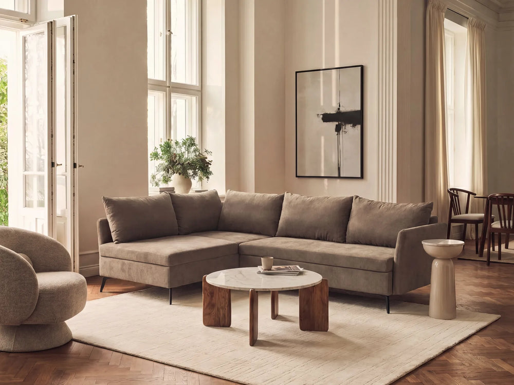 A modern L-shaped sectional sofa upholstered in taupe fabric, styled in a bright, elegant living room with a round wooden coffee table, a beige rug, and a large framed abstract print on the wall.