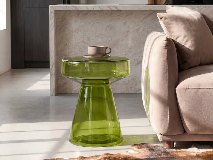 A translucent olive green glass side table with a wide round top and tapered pedestal base, styled in a modern living room next to a beige sofa.