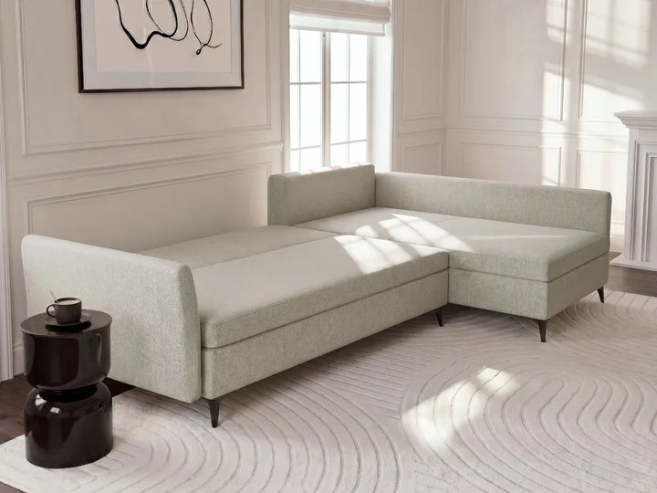 A light beige upholstered sectional sofa with a chaise lounge section, shown in a bright, modern living room with white walls, a patterned rug, and a small black side table.