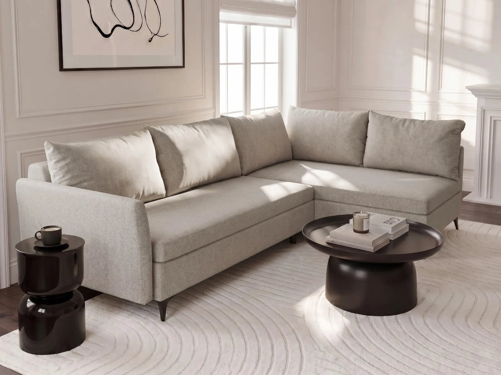 A light beige upholstered sectional sofa with matching back cushions in a bright, modern living room setting. The sofa is styled with a dark round coffee table, a black side table, and a white patterned rug.