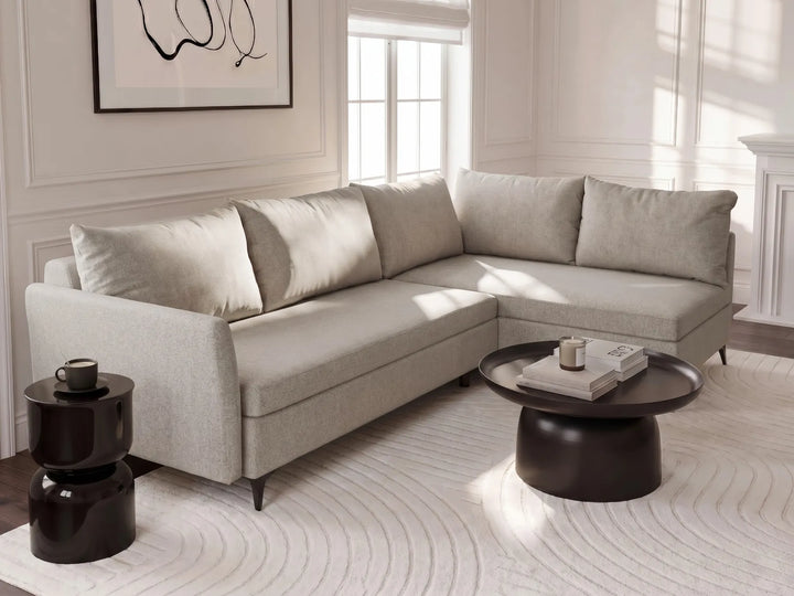 A light beige upholstered sectional sofa with matching back cushions in a bright, modern living room setting. The sofa is styled with a dark round coffee table, a black side table, and a white patterned rug.