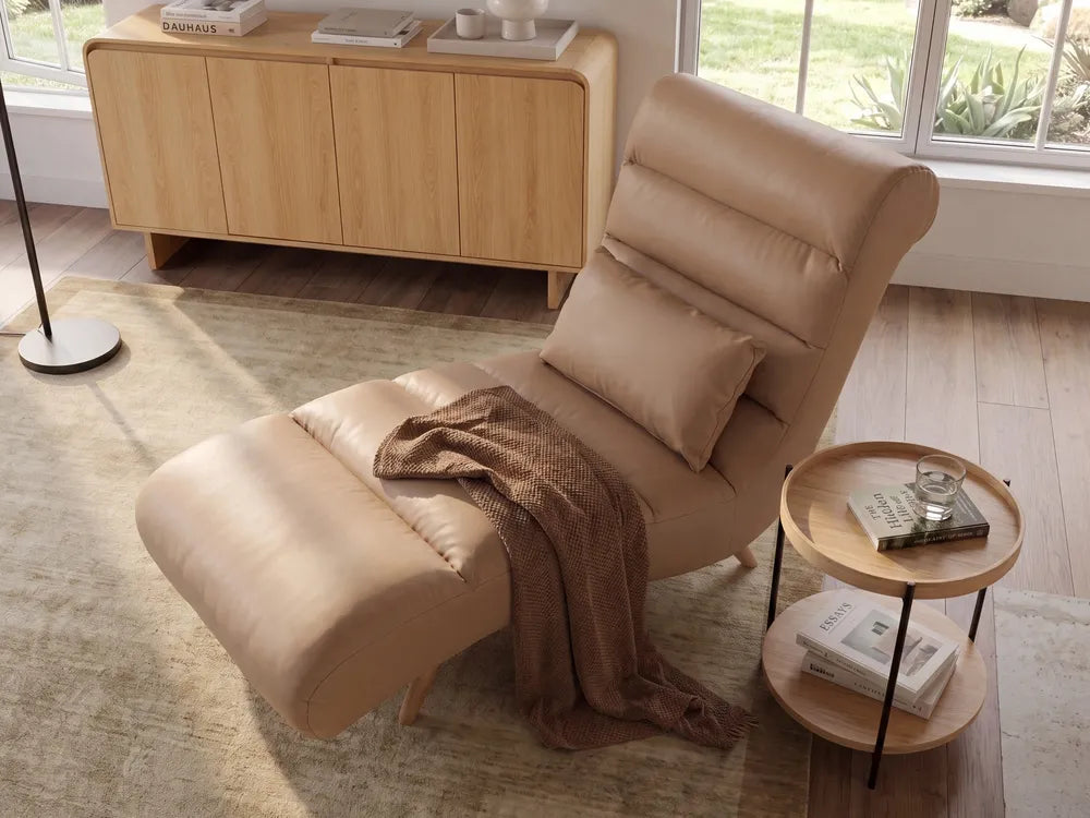 A beige leather recliner chair with an adjustable headrest and matching cushion, styled in a bright room with a wooden sideboard, window, and a small round wooden side table.