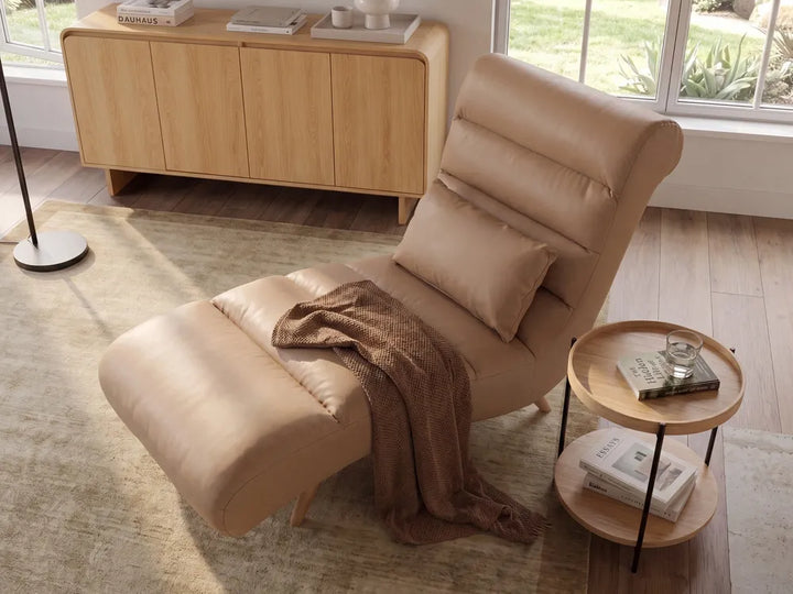 A beige leather recliner chair with an adjustable headrest and matching cushion, styled in a bright room with a wooden sideboard, window, and a small round wooden side table.