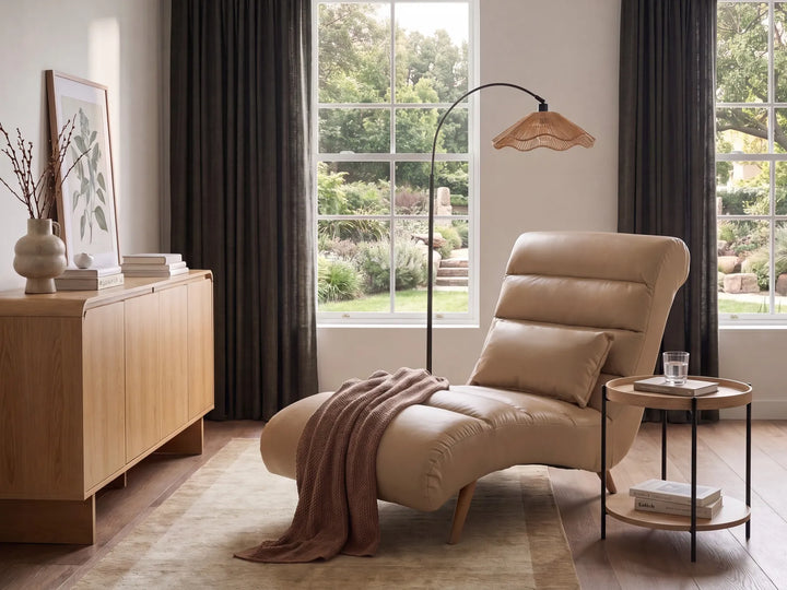 A beige leather chaise lounge chair in a modern, sunlit living room setting with dark curtains, a wooden sideboard, and a floor lamp.