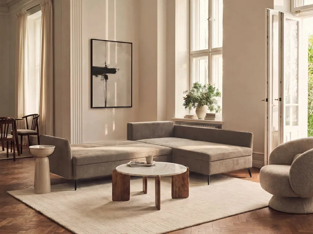 A modern living room featuring a taupe gray L-shaped sectional sofa, a round marble-top coffee table with wooden legs, a large cream rug, and warm beige walls with tall windows.