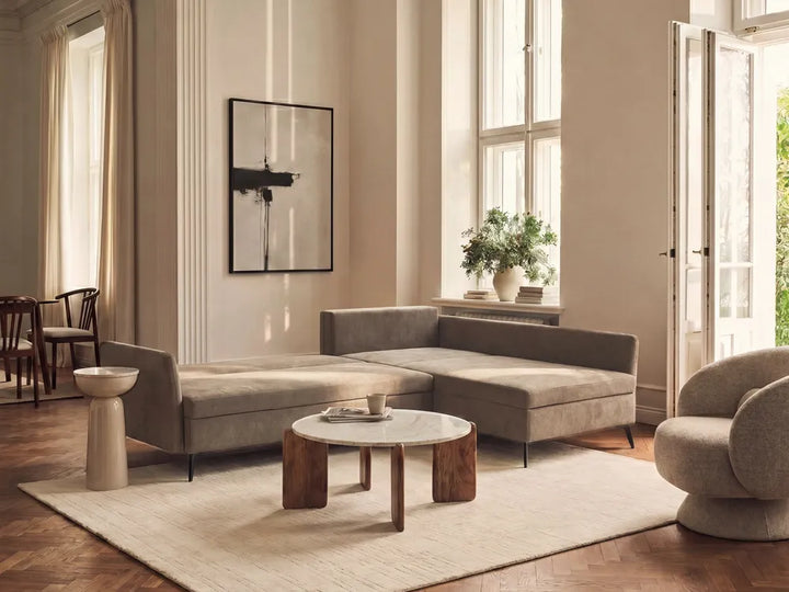A modern living room featuring a taupe gray L-shaped sectional sofa, a round marble-top coffee table with wooden legs, a large cream rug, and warm beige walls with tall windows.