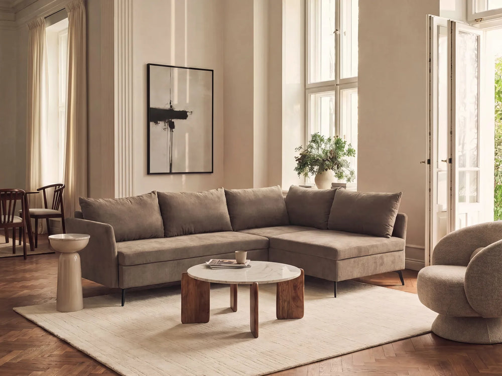 A modern L-shaped sectional sofa upholstered in a textured taupe fabric, styled in a bright, elegant living room with a round wooden coffee table, a beige rug, and warm natural light from large windows.