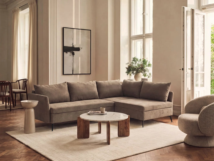 A modern L-shaped sectional sofa upholstered in a textured taupe fabric, styled in a bright, elegant living room with a round wooden coffee table, a beige rug, and warm natural light from large windows.
