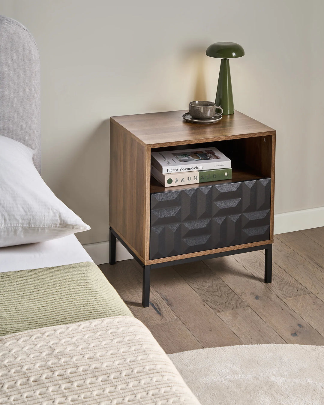Wooden nightstand with geometric design next to a bed with white pillows and green blanket.