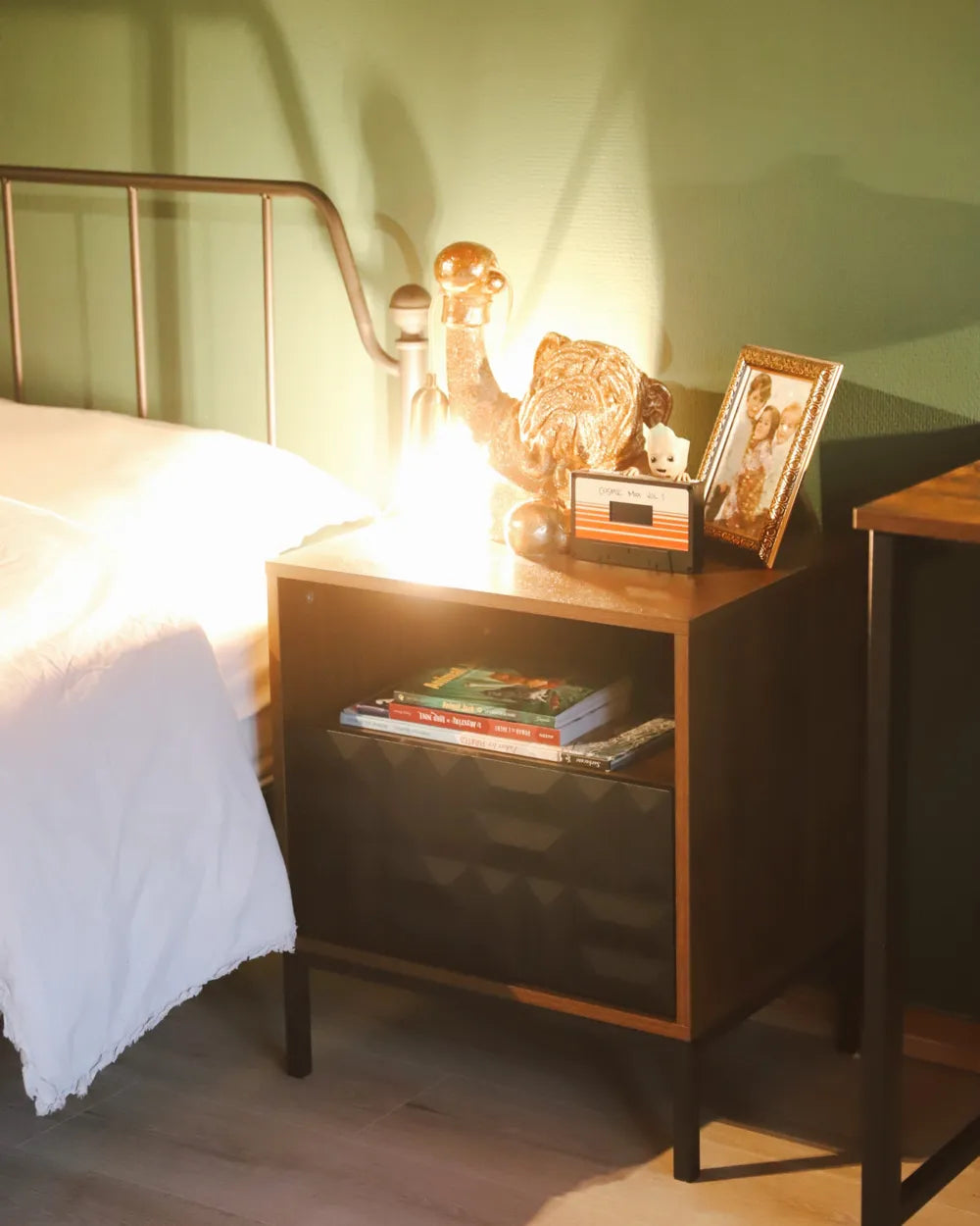 A mid-century modern wooden nightstand with a dark finish and glass-front lower cabinet, styled in a bedroom next to a bed with white bedding and a metal headboard.