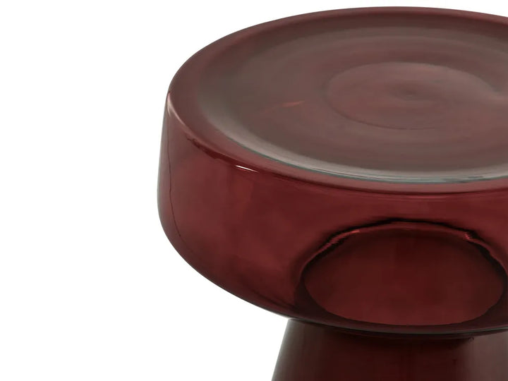 A close-up of a round, low stool or side table made from translucent burgundy glass, showing the thick circular seat and a matching pedestal base against a plain white background.