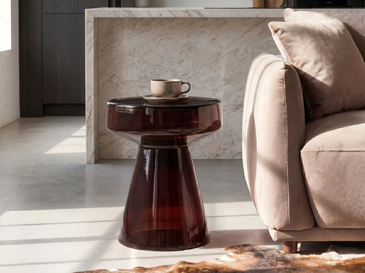 A modern sculptural side table made of translucent dark amber glass, styled in a minimalist living room next to a beige sofa.
