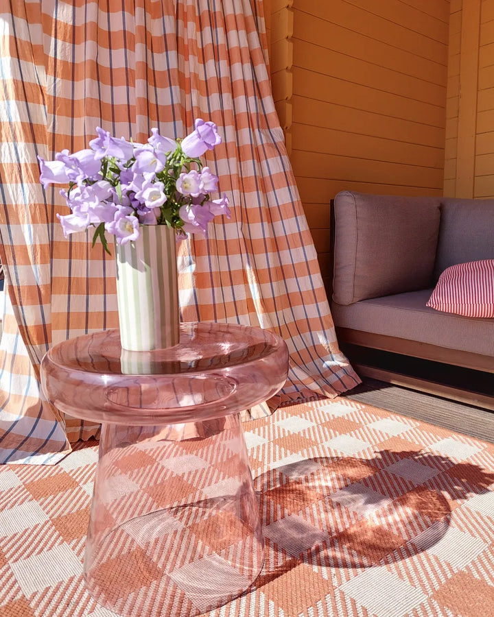 Pink table with a vase of purple flowers against a checkered curtain and wooden wall.