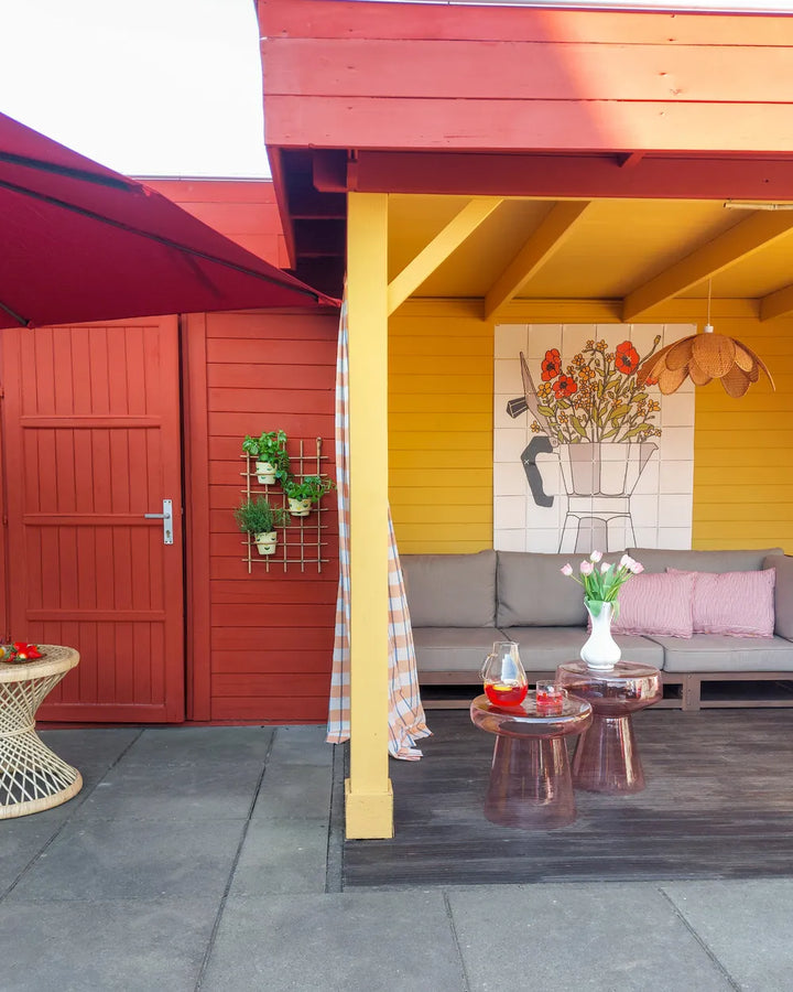 A colorful outdoor patio area with a red roof and red walls, a yellow wall, a gray sofa with pink pillows, and small round tables in the foreground.